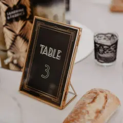 Un nom de table mariage chic, "TABLE 3", dans un cadre doré, entouré de bougies décoratives et d'une tranche de pain.