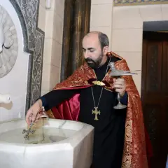 Un prêtre bénit l'eau pour un baptême arménien, un jeune garçon en costume bleu observe attentivement.