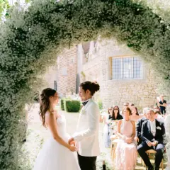 Un couple se tient sous une arche de fleurs blanches lors d'une cérémonie en plein air. L'homme, le parrain de cœur, porte un costume blanc.