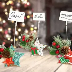 Trois étoiles décoratives de Noël, ornées de branches de sapin, de baies rouges et de pommes de pin, servent d'étiquette marque place pour "Papa", "Marie" et "Maman".