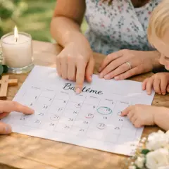 Planification du baptême : des doigts pointent des dates sur un calendrier, cherchant le samedi ou dimanche idéal. Bougie, croix et fleurs ajoutent à l'atmosphère.
