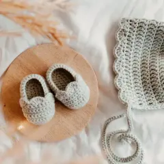 Doua nouveau-né en crochet gris, des chaussons sur un plateau en bois et un bonnet assorti, prêts pour une nouvelle aventure.