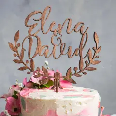 Topper gateau mariage en bois avec les prénoms "Elena & David" entourés de feuillage.