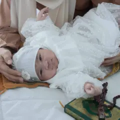 Un bébé en tenue blanche, entouré d'une mère, d'un tapis de prière, d'un Coran et de perles. Une scène évoquant les jours de naissance et leur sacrifice en Islam.
