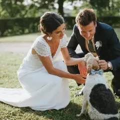Un couple heureux lors d'un mariage campagne, caressant leur chien portant un nœud papillon bleu.