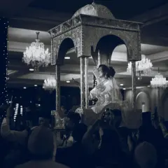 Une mariée, parée pour un baptême marocain, est portée sur un trône orné. La foule l'acclame, capturant le moment avec leurs téléphones.