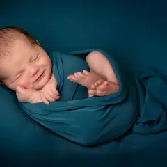 Un bébé endormi, emmailloté dans un tissu bleu canard, sourit paisiblement. Une belle image pour célébrer une naissance, un moment de joie et de gratitude, que dire pour une naissance islam.