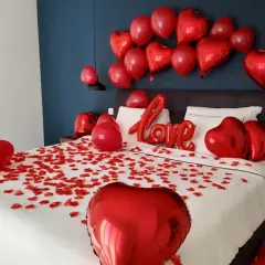 Une chambre décorée pour la Saint-Valentin, avec des pétales de rose et des ballons rouges en forme de cœur. On peut y voir une inscription "Love" en ballons.