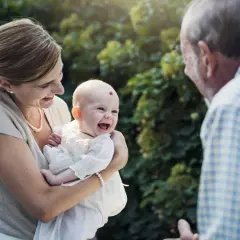 Une mère tient son bébé souriant, face à un homme. La question "faut il l'accord des 2 parents pour un bapteme civil ?" se pose.