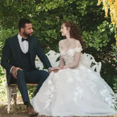 Un couple s'assoit sur un banc dans un jardin luxuriant, évoquant un mariage Bridgerton. La mariée porte une robe de bal ornée, le marié un costume sombre.