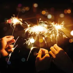 Trois mains tiennent des feux du bengale illuminant la nuit. Des étincelles dorées dansent dans l'obscurité, créant une atmosphère festive.
