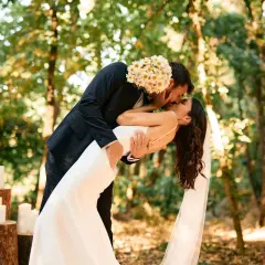 Un couple s'embrasse passionnément lors de leur mariage en forêt. Le marié tient la mariée, dont la robe blanche fluide contraste avec son costume sombre. Ce moment magique illustre parfaitement les prestations mariage.