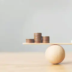Figurines de mariés en équilibre sur une balance avec des pièces. Une métaphore visuelle pour comment préparer un mariage et gérer son budget.