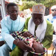 Cérémonie de baptême au Sénégal. Un bébé est tenu par deux hommes en tenue traditionnelle, entourés de fidèles.