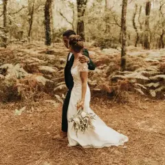 Un couple se promène dans une forêt, la mariée porte une robe blanche et tient un bouquet. Un mariage DIY plein de charme.