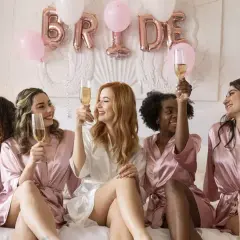 Des amies célèbrent un EVJF, c'est quoi cette fête ? Elles portent des peignoirs en satin et trinquent avec du champagne, des ballons "BRIDE" en arrière-plan.
