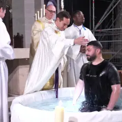 Cérémonie de baptême adulte dans une église. Un homme en t-shirt noir est immergé dans l'eau d'une cuve, entouré de religieux en soutanes blanches.