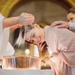 Une jeune fille souriante se fait baptiser à 16 ans. L'eau bénite coule sur sa tête, un moment émouvant.