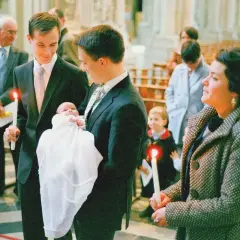Le parrain tient sa nièce, un bébé en habit blanc, lors d'une cérémonie religieuse. Une bougie allumée est tenue par un autre homme.