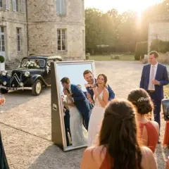 Un couple de mariés s'amuse devant un miroir, entouré d'invités. Une voiture ancienne décorée de fleurs est garée derrière eux. Une belle scène de tradition mariage.