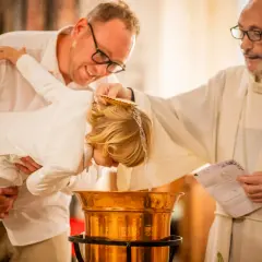 Cérémonie de baptême : un prêtre verse de l'eau sur la tête d'un enfant tenu par son père.