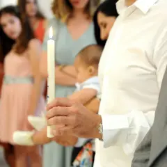 Un homme tient une bougie allumée, prêt à devenir parrain catholique. Un bébé est porté à proximité.