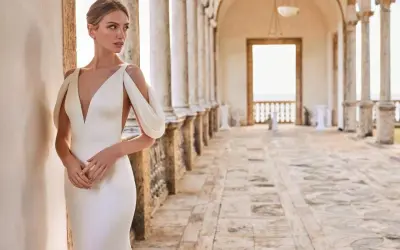 Une mariée essaie une robe élégante, avec un décolleté plongeant et des manches drapées, dans un cadre architectural lumineux.