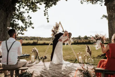Un couple s'embrasse lors d'une **cérémonie laïque extérieur**. Un arche triangulaire décoré de fleurs et de plumes se dresse derrière eux.