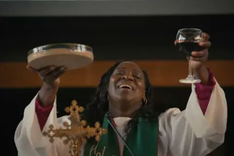 Une femme, peut-être une marraine de substitution, célèbre la communion en levant une coupe de vin et une patène.