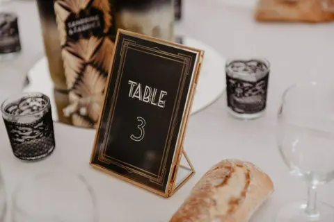 Un nom de table mariage chic, "TABLE 3", dans un cadre doré, entouré de bougies décoratives et d'une tranche de pain.