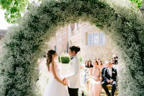 Un couple se tient sous une arche de fleurs blanches lors d'une cérémonie en plein air. L'homme, le parrain de cœur, porte un costume blanc.