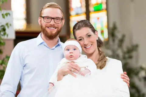 Un couple souriant pose avec leur bébé lors d'un baptême. La question "peut-on être marraine à 11 ans ?" résonne, mais ici, c'est une famille unie.