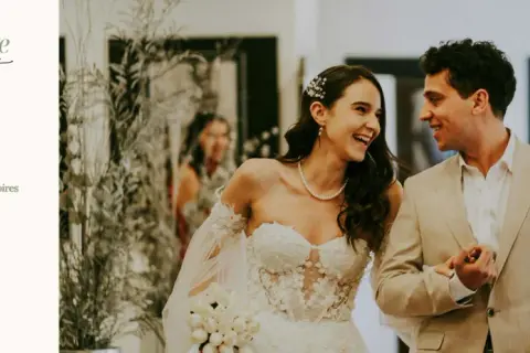 Un couple souriant, une mariée en robe blanche et un marié en costume beige, planifiant leur mariage sur une liste de tâches.