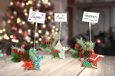 Trois étoiles décoratives de Noël, ornées de branches de sapin, de baies rouges et de pommes de pin, servent d'étiquette marque place pour "Papa", "Marie" et "Maman".