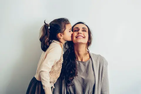 Une petite fille embrasse sa mère sur la joue. Elle pense : "je ne sais pas qui choisir comme marraine", mais l'amour est évident.