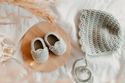 Doua nouveau-né en crochet gris, des chaussons sur un plateau en bois et un bonnet assorti, prêts pour une nouvelle aventure.