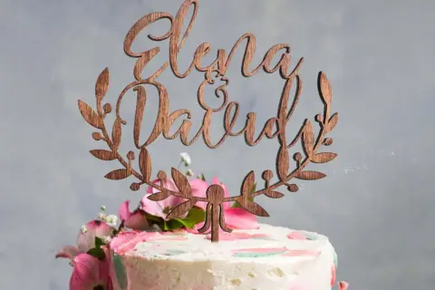 Topper gateau mariage en bois avec les prénoms "Elena & David" entourés de feuillage.