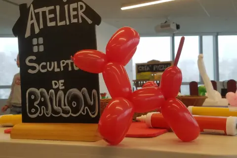 Un chien en sculpture ballon rouge, à côté d'un panneau "Atelier Sculpture de Ballon". Facile à réaliser !