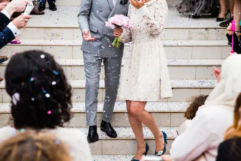 Un couple de mariés, sous une pluie de confettis, s'apprête à lancer la chanson d'ouverture de bal. La mariée tient un bouquet de roses roses.