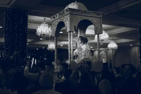 Une mariée, parée pour un baptême marocain, est portée sur un trône orné. La foule l'acclame, capturant le moment avec leurs téléphones.