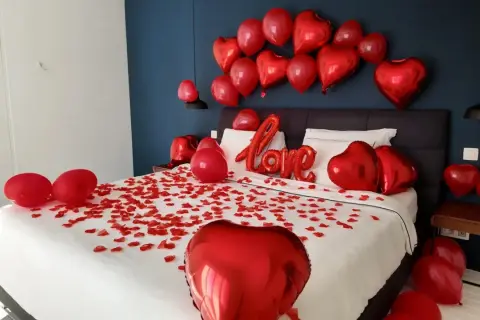 Une chambre décorée pour la Saint-Valentin, avec des pétales de rose et des ballons rouges en forme de cœur. On peut y voir une inscription "Love" en ballons.