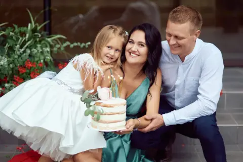 Famille souriante autour d'un gâteau pour un baptême civil ou religieux. La petite fille porte une robe blanche ornée de plumes.