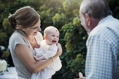 Une mère tient son bébé souriant, face à un homme. La question "faut il l'accord des 2 parents pour un bapteme civil ?" se pose.