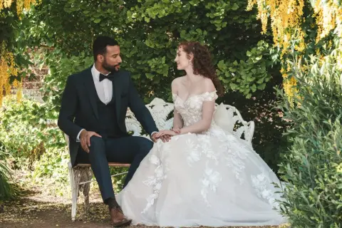 Un couple s'assoit sur un banc dans un jardin luxuriant, évoquant un mariage Bridgerton. La mariée porte une robe de bal ornée, le marié un costume sombre.