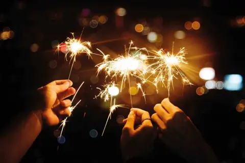 Trois mains tiennent des feux du bengale illuminant la nuit. Des étincelles dorées dansent dans l'obscurité, créant une atmosphère festive.