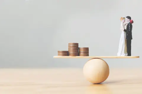 Figurines de mariés en équilibre sur une balance avec des pièces. Une métaphore visuelle pour comment préparer un mariage et gérer son budget.