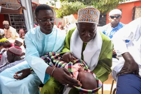Cérémonie de baptême au Sénégal. Un bébé est tenu par deux hommes en tenue traditionnelle, entourés de fidèles.