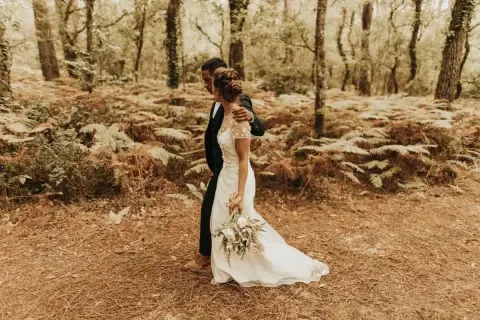 Un couple se promène dans une forêt, la mariée porte une robe blanche et tient un bouquet. Un mariage DIY plein de charme.