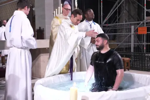 Cérémonie de baptême adulte dans une église. Un homme en t-shirt noir est immergé dans l'eau d'une cuve, entouré de religieux en soutanes blanches.