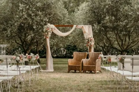 Un bel exemple de cérémonie laïque en plein air, avec un arche fleuri, des chaises blanches et deux fauteuils en rotin sous les oliviers.